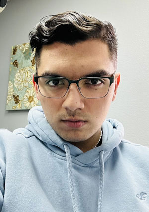 A man in a blue sweatshirt and glasses, with a beard and mustache, is looking directly at the camera.