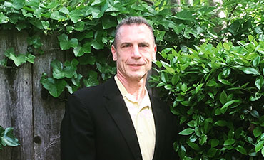 The image shows a man in a suit standing outdoors, positioned behind a hedge with a blurred background.
