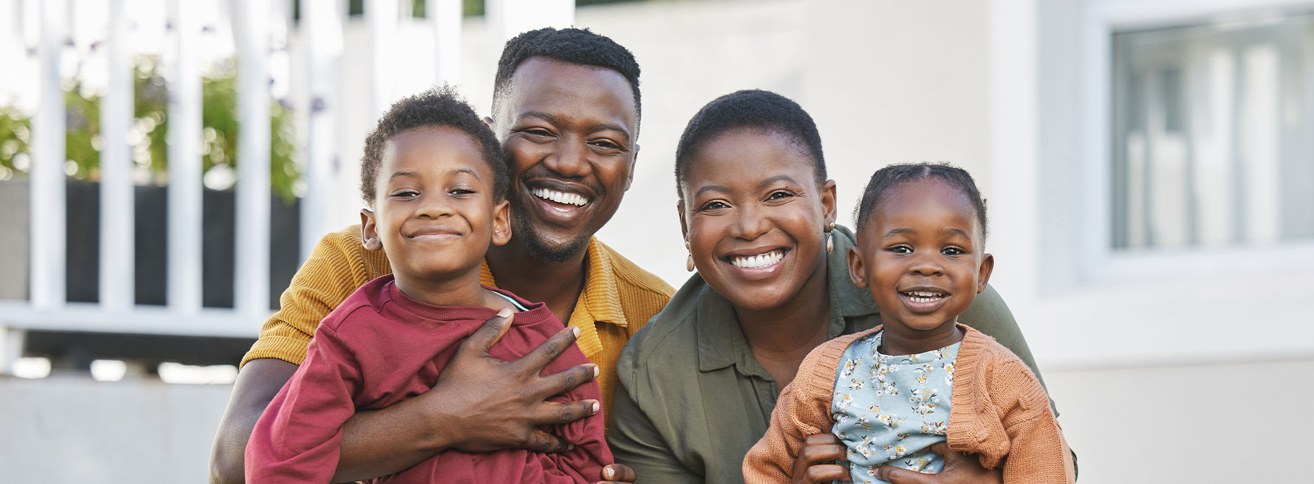 The image is a photograph of a family posing together outdoors, with the father holding two small children and the mother standing behind them.
